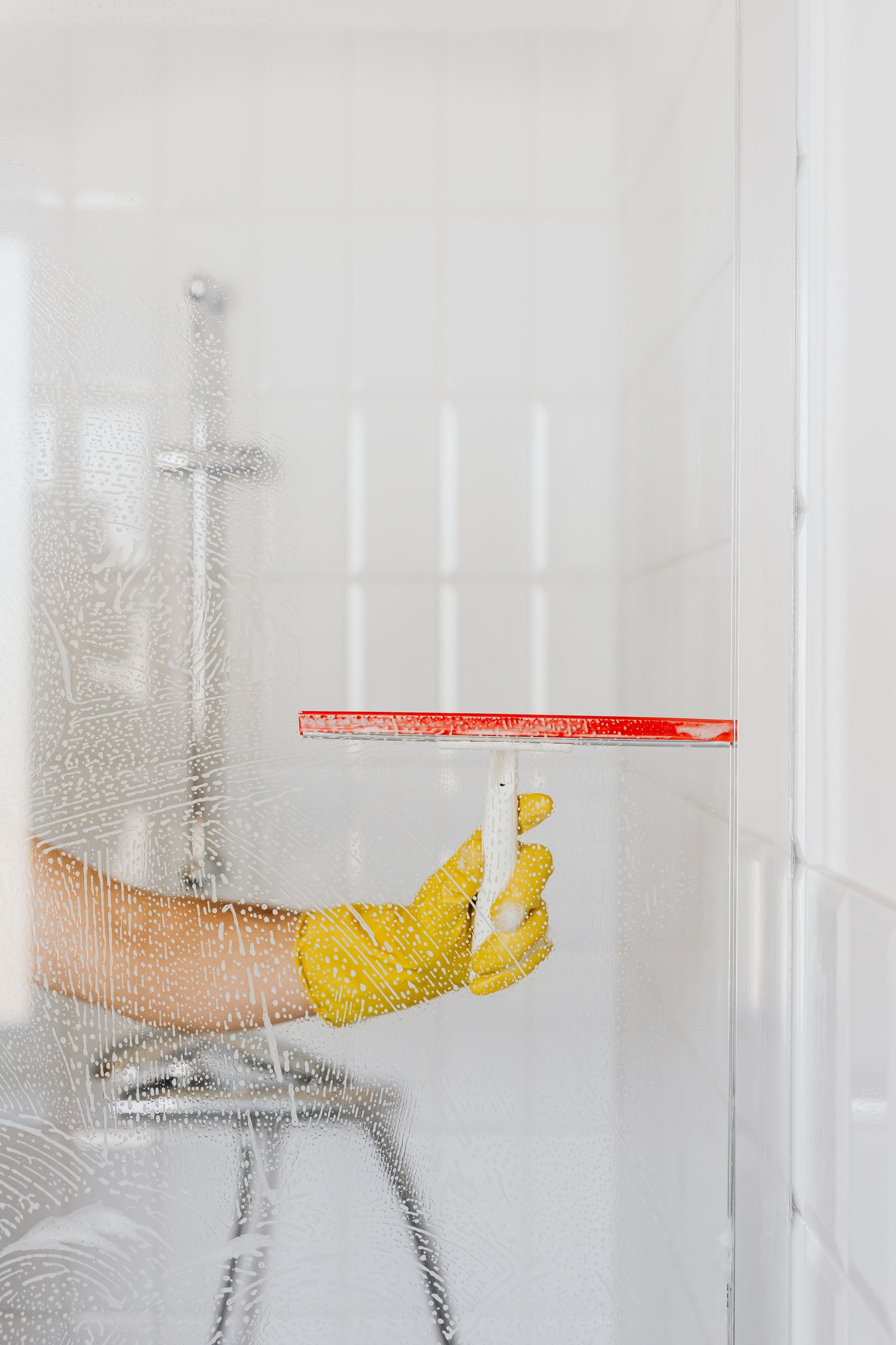 Crop anonymous person in yellow rubber glove cleaning glass shower unit with glass wiper and detergent against white tile wall